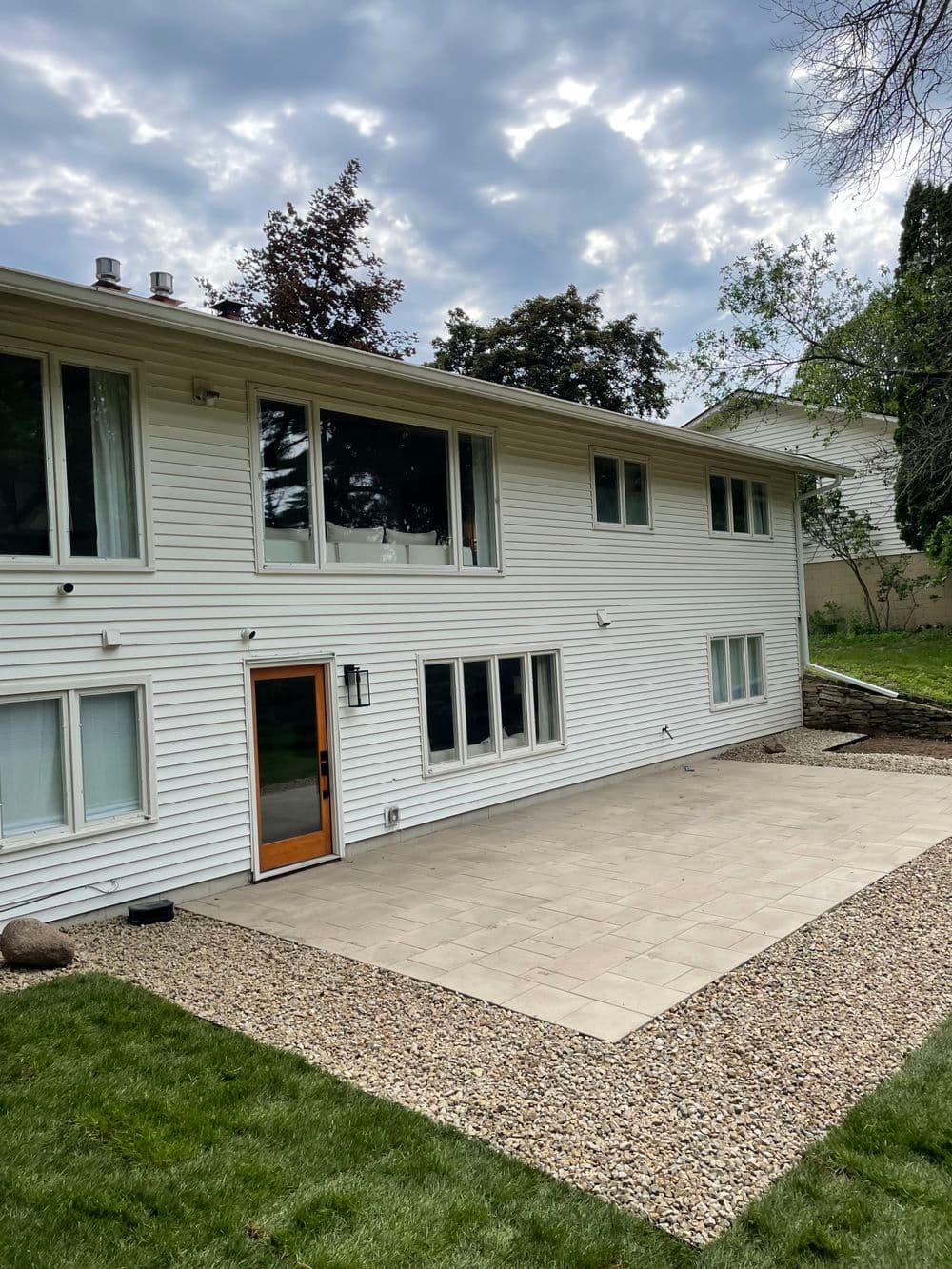 Modern home exterior with large windows, stone patio, and landscaped yard under a cloudy sky.
