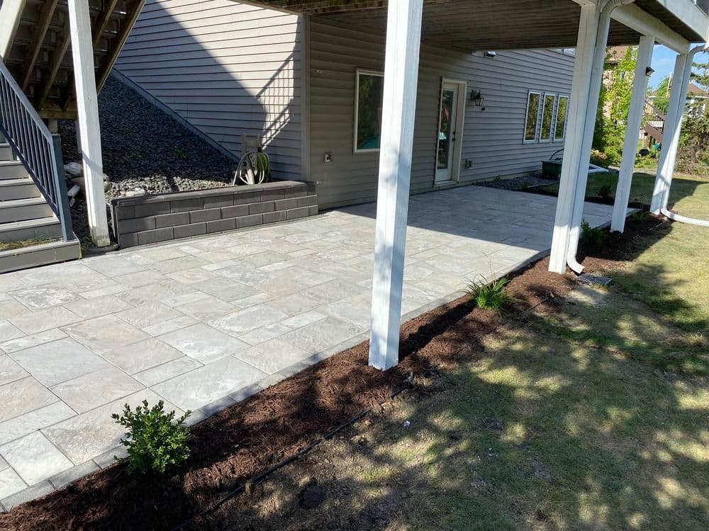 Modern patio with stone pavers, landscaped edges, and a shaded overhang under a house.