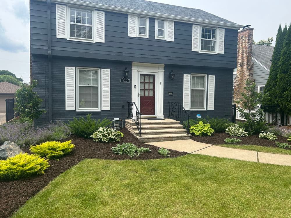 Dark gray traditional house with white shutters, lush landscaping, and stone pathway.