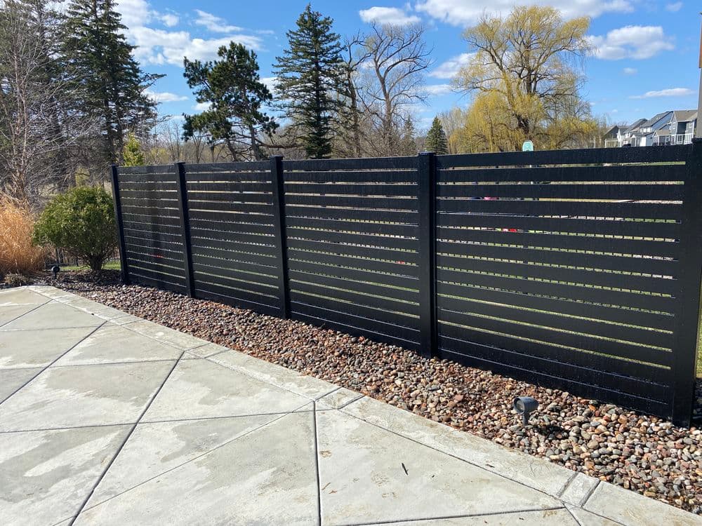 Modern black horizontal slat fence in a landscaped yard with trees and blue sky.
