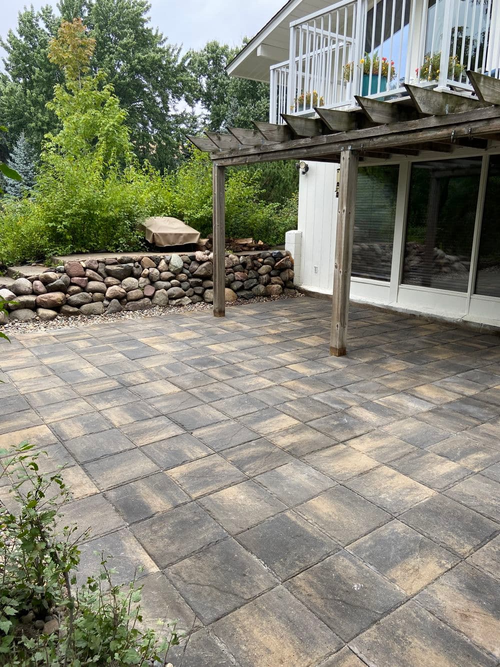 Patio area with stone paving, wooden pergola, and greenery near a house.