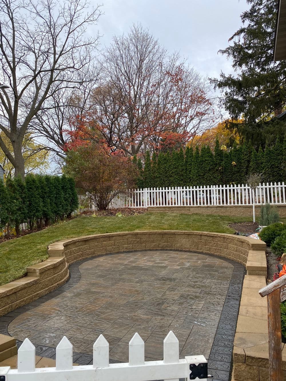 Stylish circular patio with stone surface and autumn trees, surrounded by greenery and white fence.