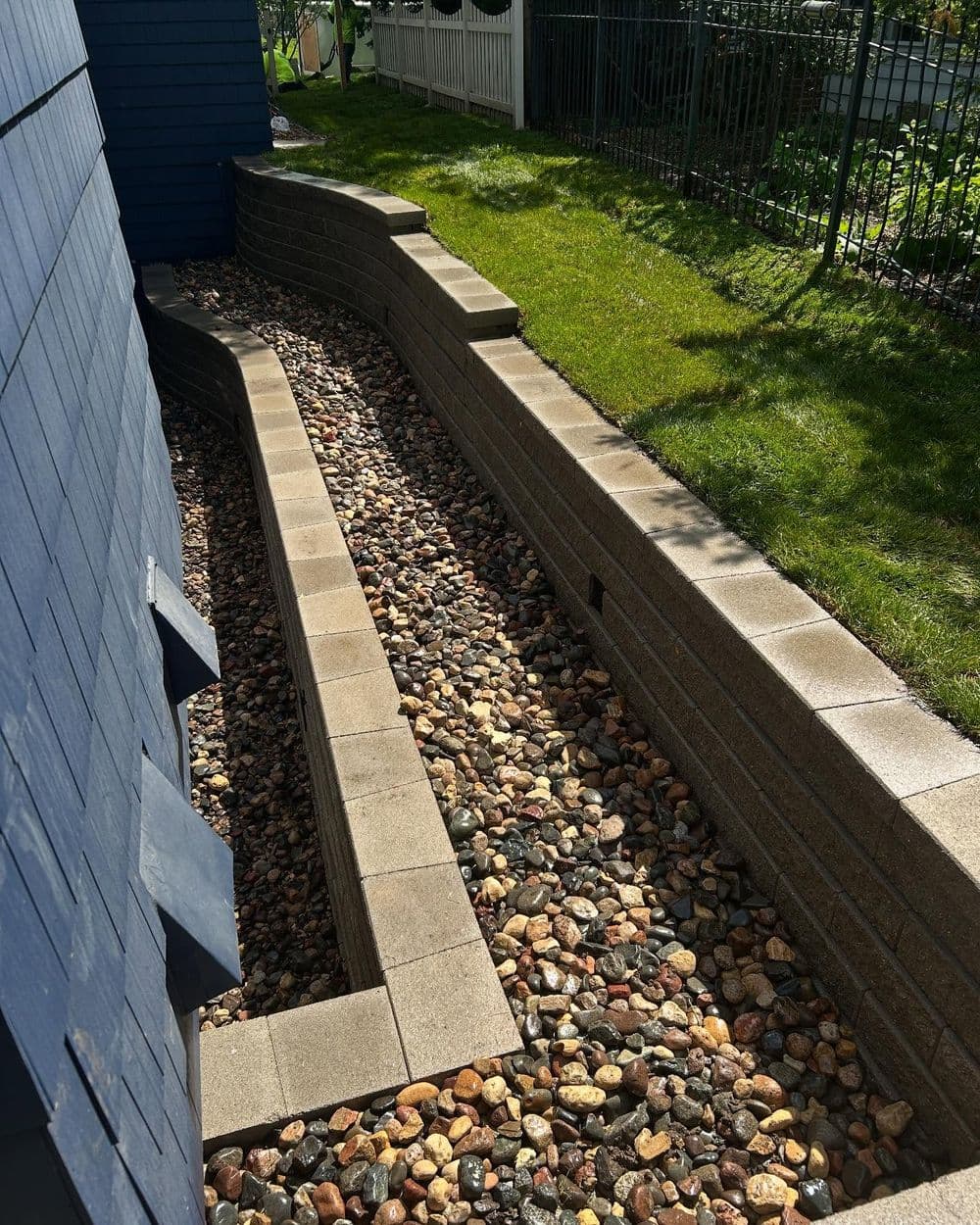 Landscaped drainage area with stone pathway and retaining walls beside a blue house.