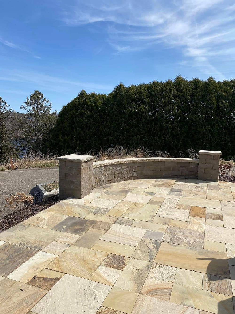 Curved stone patio with light-colored paving stones and lush green hedges in the background.
