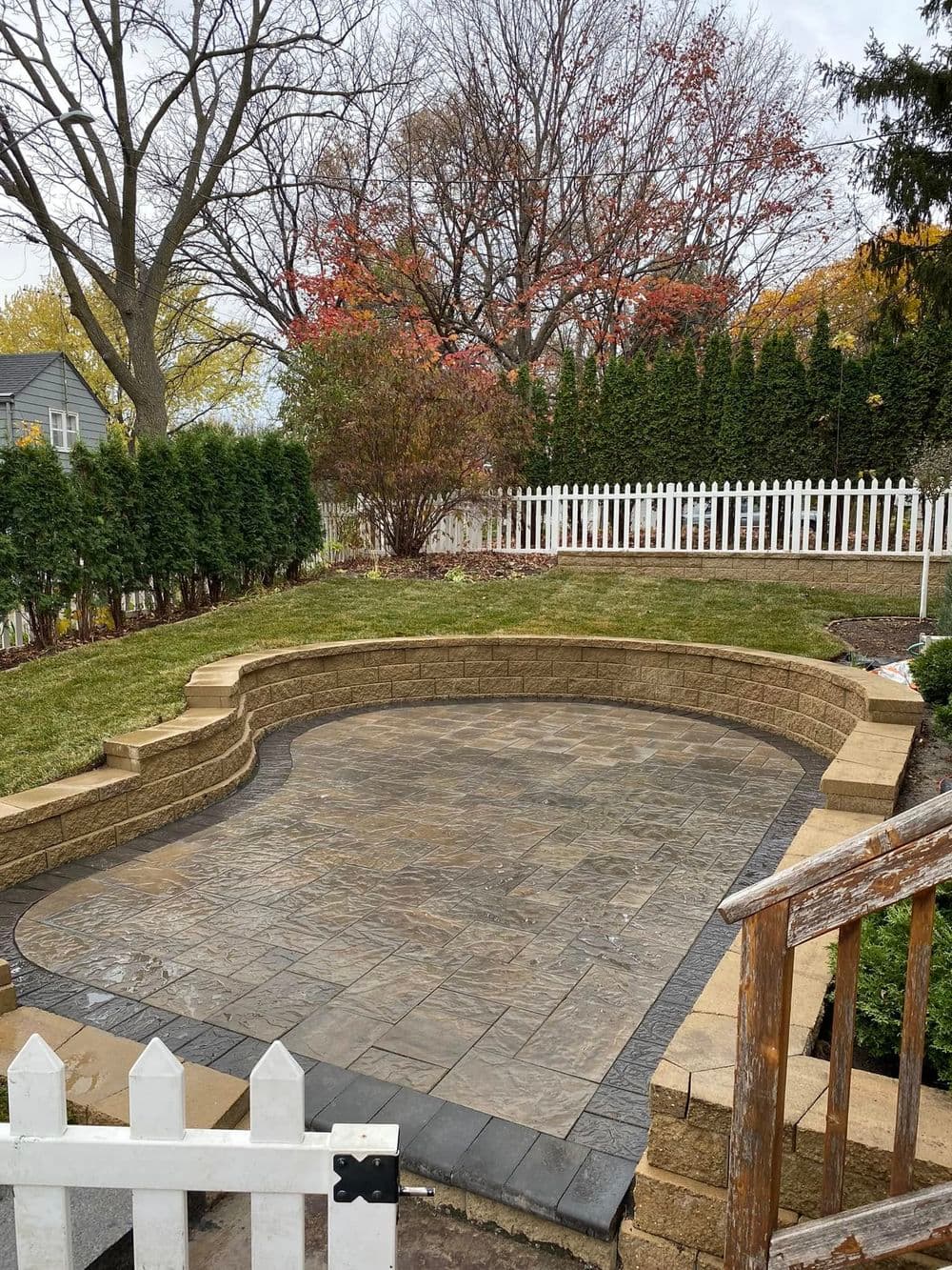 Landscaped backyard featuring a stone patio, greenery, and a white picket fence.
